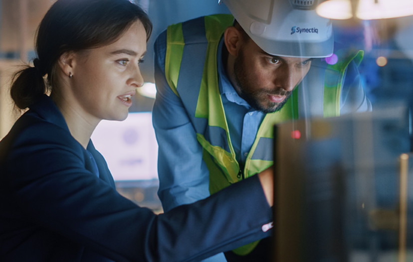 A woman in a business suit is showing a man dressed in safety equipment something on a flat monitor.
